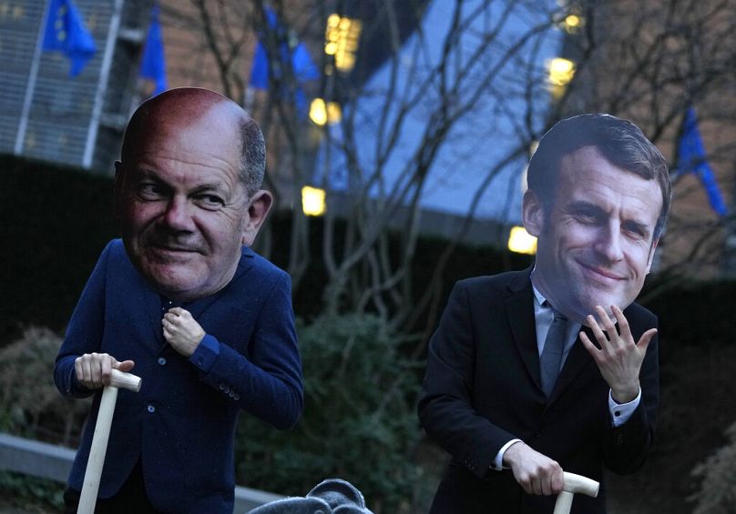 Aktywiści w maskach kanclerza Niemiec Olafa Scholza i prezydenta Francji Emmanuela Macrona trzymają łopaty przed siedzibą UE w Brukseli, luty 2022 r.