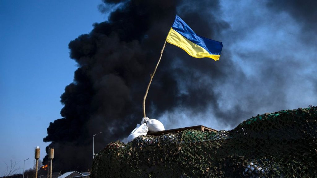 A Ukrainian flag is seen on top of a checkpoint as black smoke rises from a fuel storage of the Ukrainian army following a Russian attack, on the outskirts of Kyiv, Ukraine, F