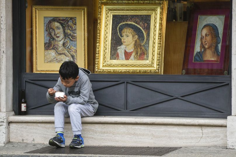 Dziecko je lody, siedząc przy oknie sklepu sprzedającego przedmioty religijne w Rzymie, styczeń 2023 r