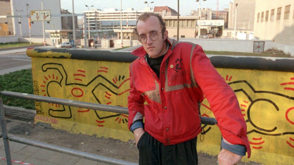 Why is an AI altered Keith Haring painting sparking outrage?  Pictured here: Haring stands in front of part of the Berlin Wall that he painted in Berlin - 1986