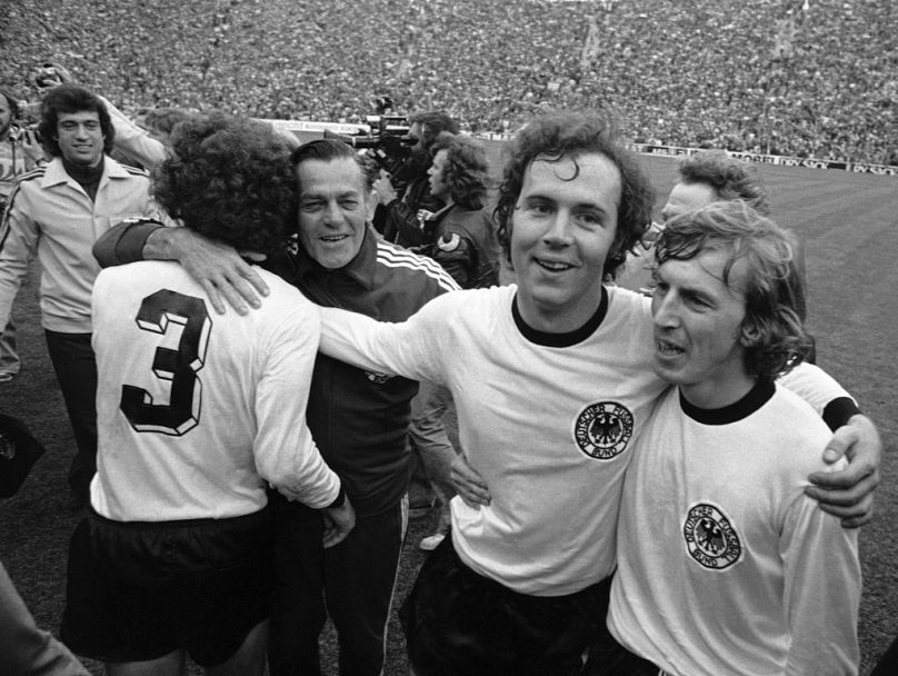Kapitan reprezentacji Niemiec Zachodnich w piłce nożnej Franz Beckenbauer, drugi od prawej, obejmuje swojego kolegę z drużyny, napastnika Juergena Grabowskiego, spacerując po stadionie olimpijskim.