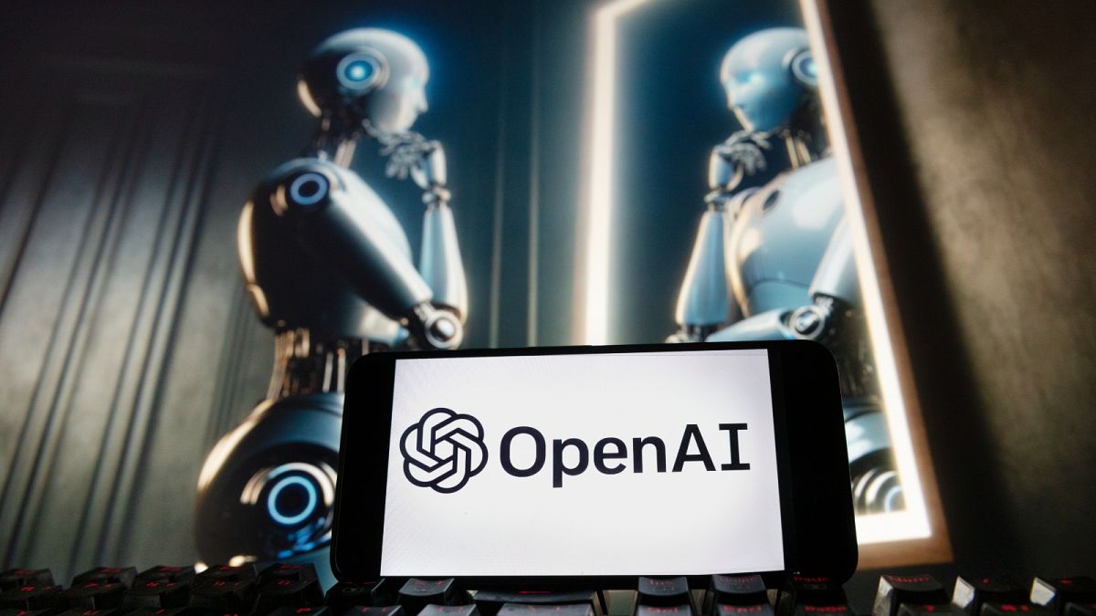 The OpenAI logo is seen displayed on a cell phone with an image on a computer monitor generated by ChatGPT