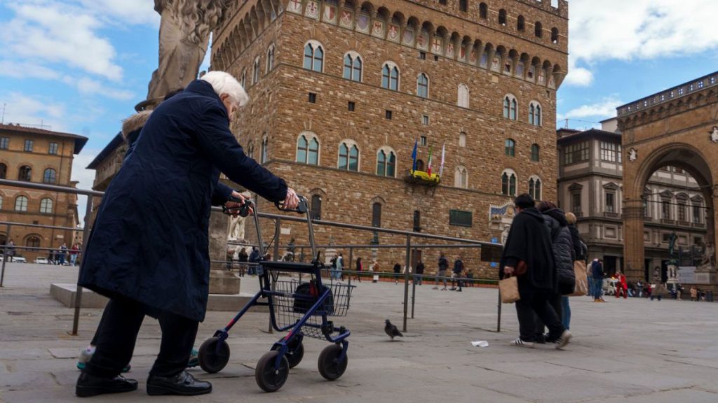 An elderly woman uses a walking frame to ambulate in front of the 14th-century town hall