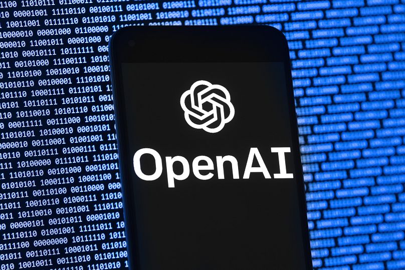 Logo OpenAI pojawia się na telefonie komórkowym przed ekranem komputera z losowymi danymi binarnymi, czwartek, 9 marca 2023