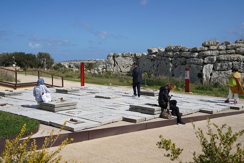 Zwiedzający siedzący i stojący na instalacji Ibrahima Mahamy „Ogród blizn” z parkiem archeologicznym Ġgantija w tle.