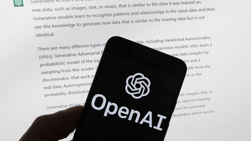 The OpenAI logo is seen on a mobile phone in front of a computer screen displaying output from ChatGPT, March 21, 2023.