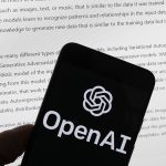 The OpenAI logo is seen on a mobile phone in front of a computer screen displaying output from ChatGPT, March 21, 2023.