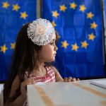 A little girl waits for her mother to vote, backdropped by voting booths with curtains depicting the European Union flag in Baleni, Romania, in 2019.