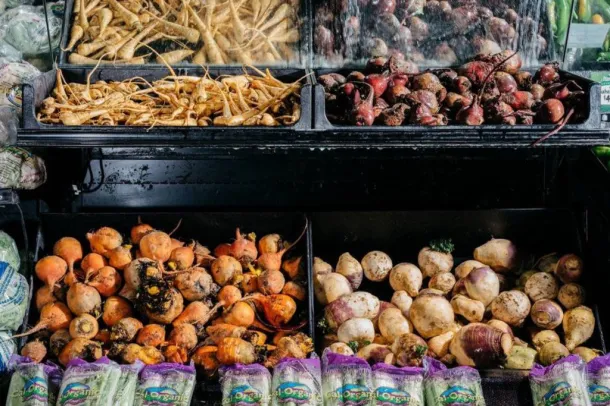Świeże warzywa korzeniowe, w tym pasternak, rzepa i buraki, są wystawiane w pojemnikach na rynku. Na pierwszym planie znajdują się torby z zieleniną Cal Organic.