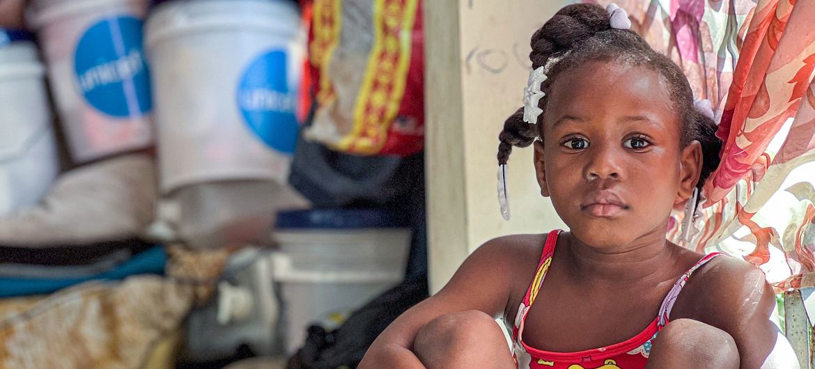Młoda Haitańska dziewczyna siedzi w zatłoczonym schronisku po ucieczce przed grupową przemocą. W tle widać zaopatrzenie UNICEF.