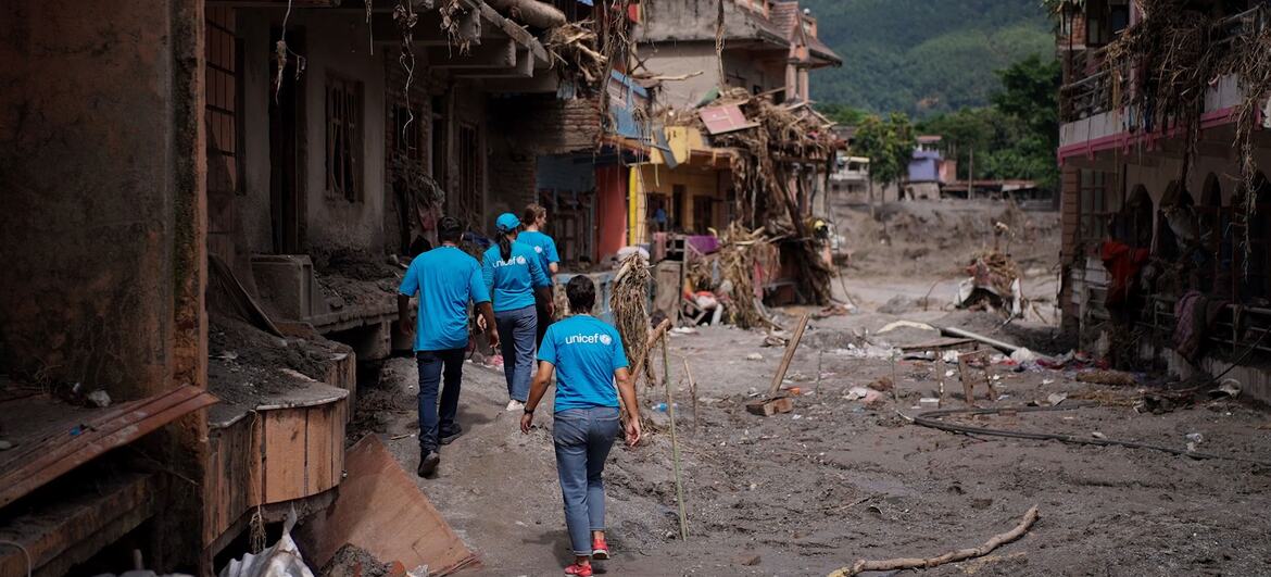 Pracownicy UNICEF w niebieskich koszulach idą zalaną ulicą w Nepalu, otoczoną uszkodzonymi budynkami i gruzem.