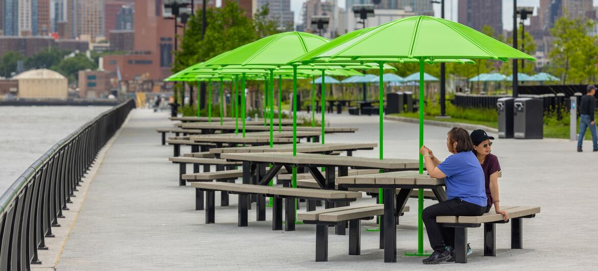 Dwie kobiety siedzą przy stołach piknikowych pod jasnozielonymi parasolami na nadmorskiej promenadzie wzdłuż East River, na tle panoramy Manhattanu.