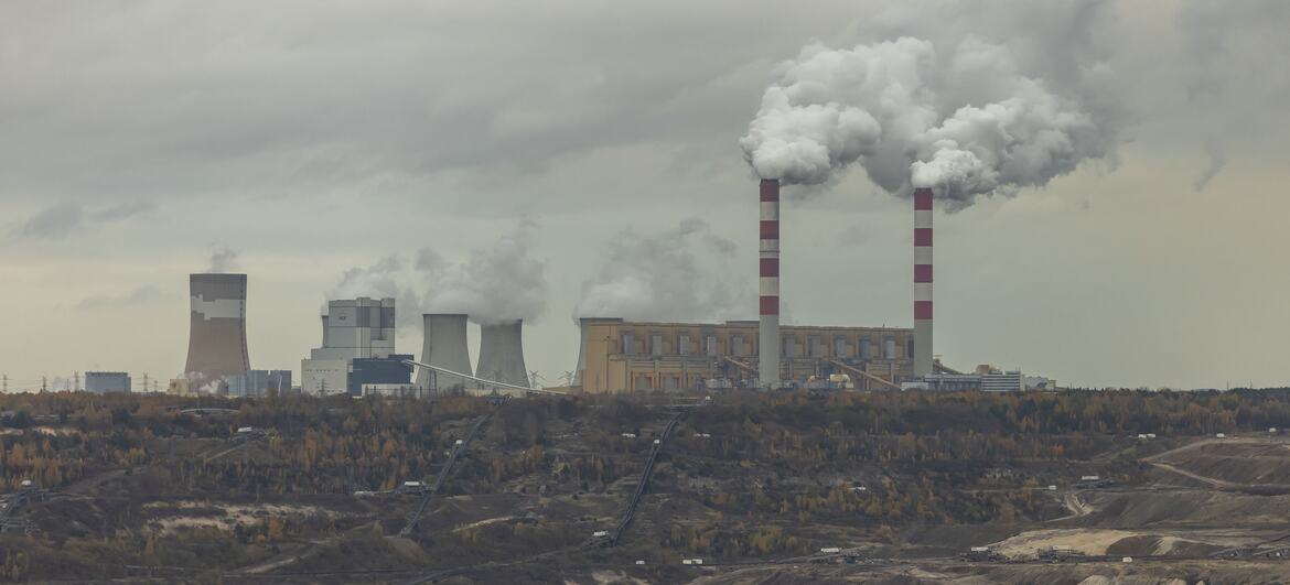 Szeroki widok na elektrownię węglową Bełchatów w Złobnicy w Polsce, ukazujący jej wieże chłodnicze i wysokie kominy w paski, uwalniające kłęby dymu nad rozległym krajobrazem górnictwa odkrywkowego pod zachmurzonym niebem.