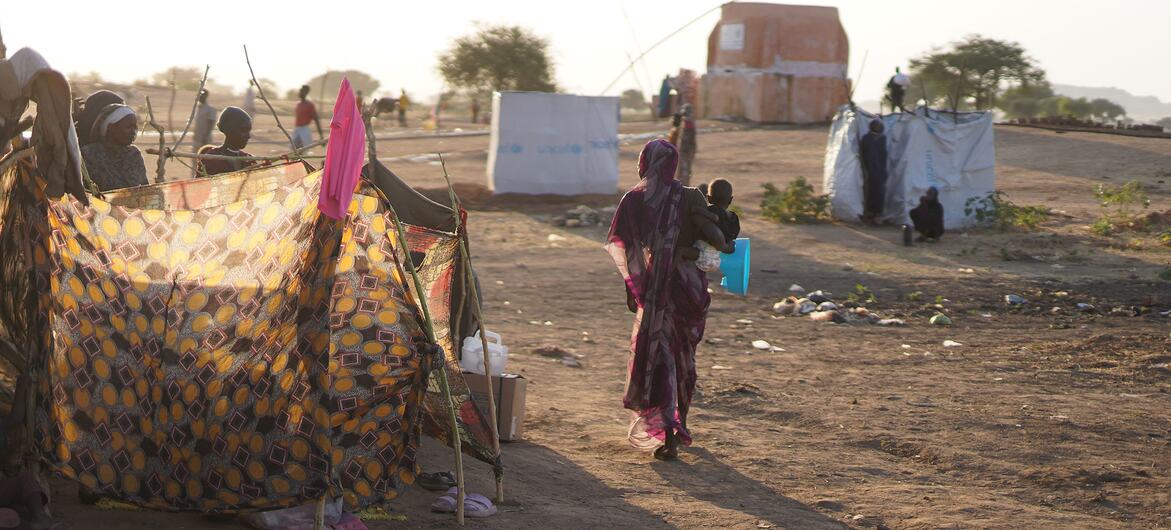 Matka przechodzi przez obóz dla przesiedleńców w Tawila w Darfurze Północnym w Sudanie, niosąc dziecko obok prowizorycznych schronisk z tkaniny i plandek z logo UNICEF na suchej ziemi.