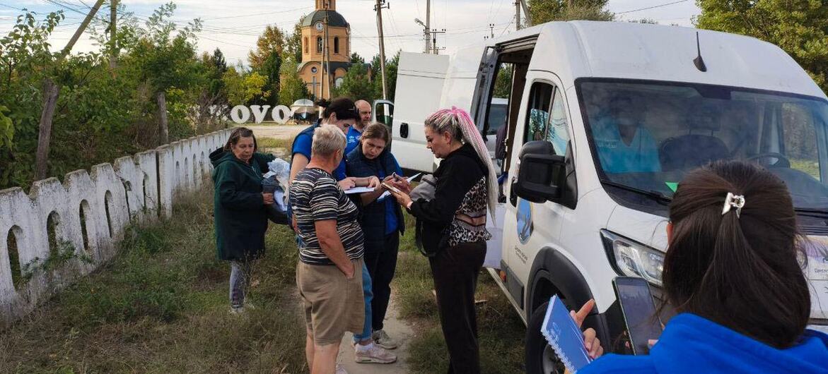 Pracownicy terenowi UN OCHA w niebieskich koszulach rozmawiają z ukraińskimi kobietami i mężczyznami obok furgonetki z białą pomocą na wiejskim poboczu, trzymając dokumenty i tablet, na tle wieży kościelnej i płotu.
